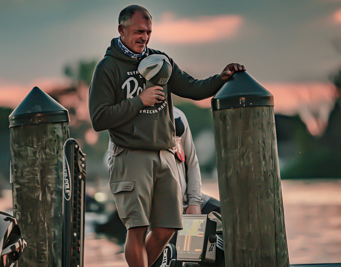 A man holds his cap over his heart on a dock at sunrise.