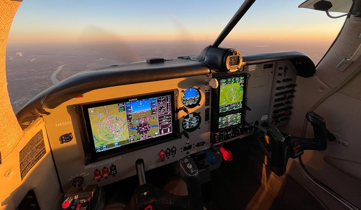 A cockpit is displayed with Garmin avionics on board while a general aircraft is mid-flight. A body of water and land can also be seen.