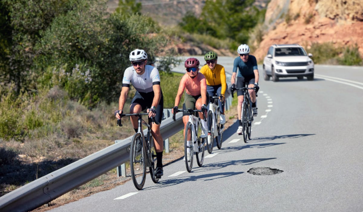 A group of cyclists ride in a single file line in a bike lane on a street as a vehicle approaches from behind.