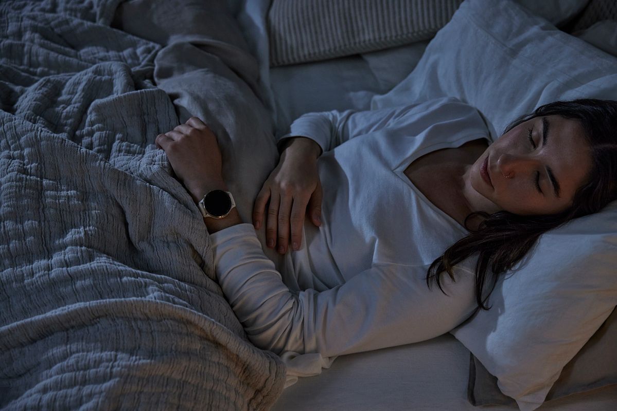 A woman in a white shirt, wearing a Garmin Venu 3, sleeps in a dark room.