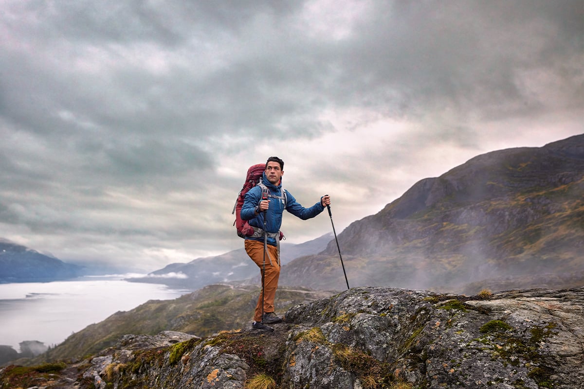 A man in a blue jacket, orange pants and red backpack with a Garmin inReach Mini 2 clipped to it hikes with mountains in the background.