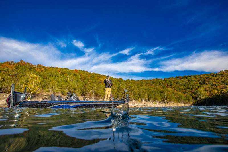 An angler located on a boat in the background appears to have caught a fish as a splash of water is visible in the foreground. The angler is alone on his boat on a lake with trees and blue skies also visible.