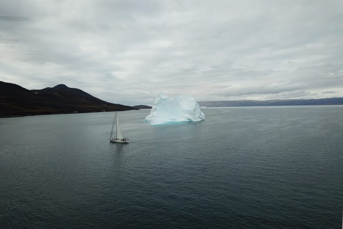 The Dogbark! sailboat sails the dark blue water of the Northwest Passage, with an iceberg in the background.