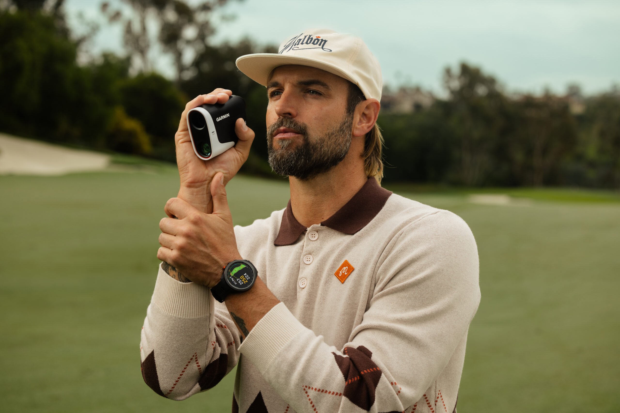 A man in a cream and brown sweater uses the Garmin Approach Z30 golf rangefinder.