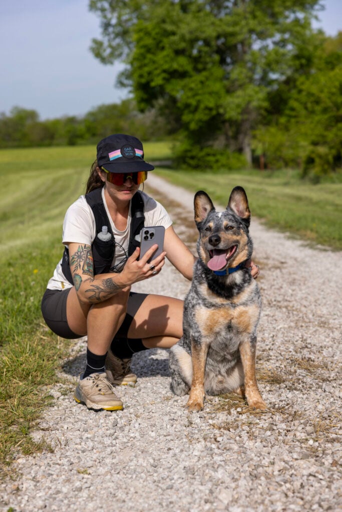 A woman in a white T-shirt and dark shorts checks the Alpha app on her phone, squatting next to her dog, which wears the Alpha LTE GPS dog tracker.