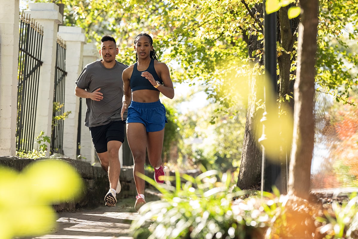 A man and a woman, wearing Garmin Forerunner smartwatches, run down a neighborhood street.
