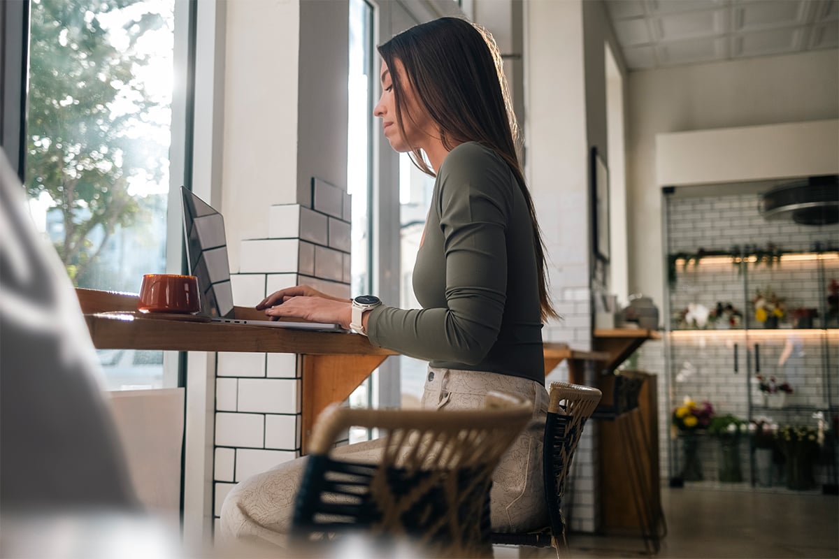 A woman in a green long sleeve shirt sits at a table while working on a laptop.