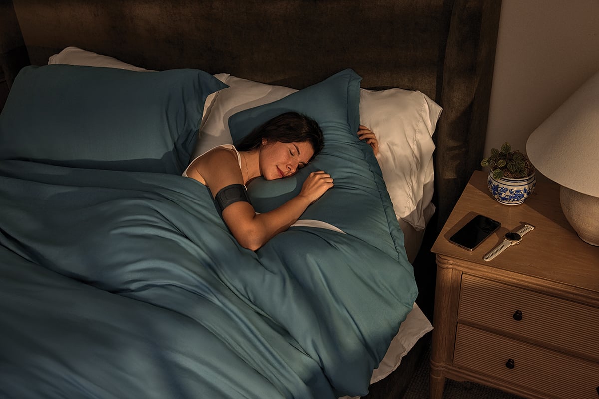 A woman wearing the Garmin Index Sleep Monitor sleeps, with her Garmin smartwatch and phone placed on her beside table.