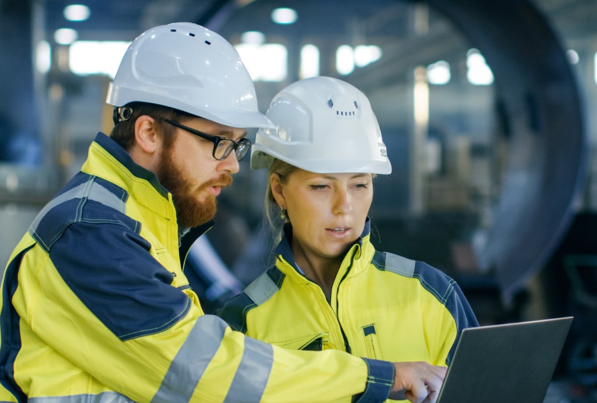 Two people wearing white hard hats look at a screen at work.