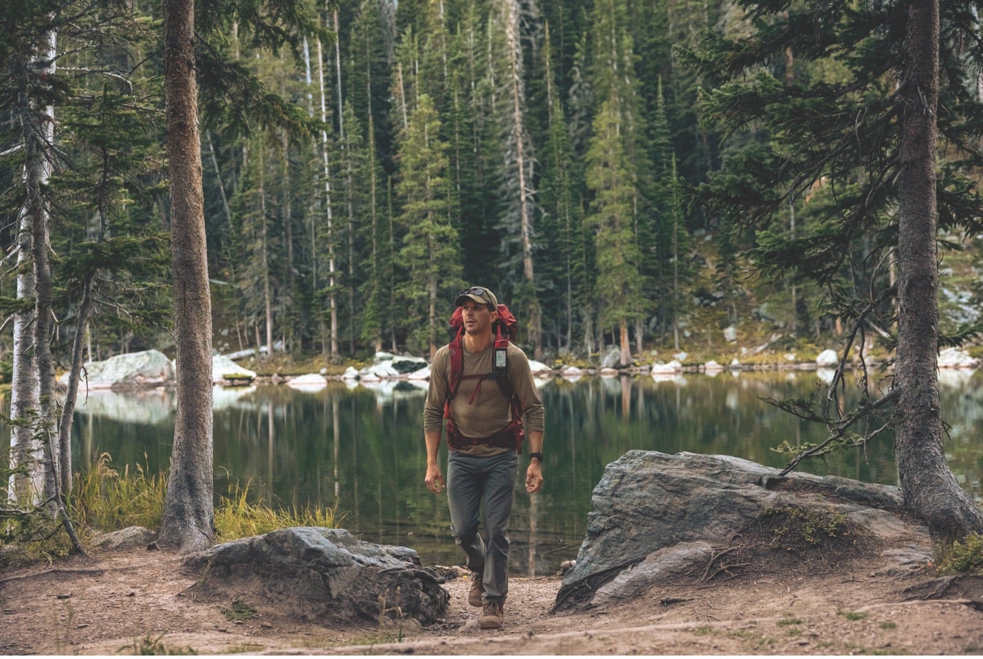 A man hiking through the forest with a lake and tall trees behind him.