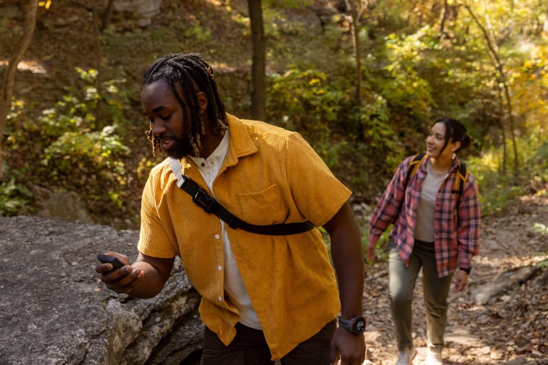 A man glances down at his device while hiking with a female a few steps behind him.