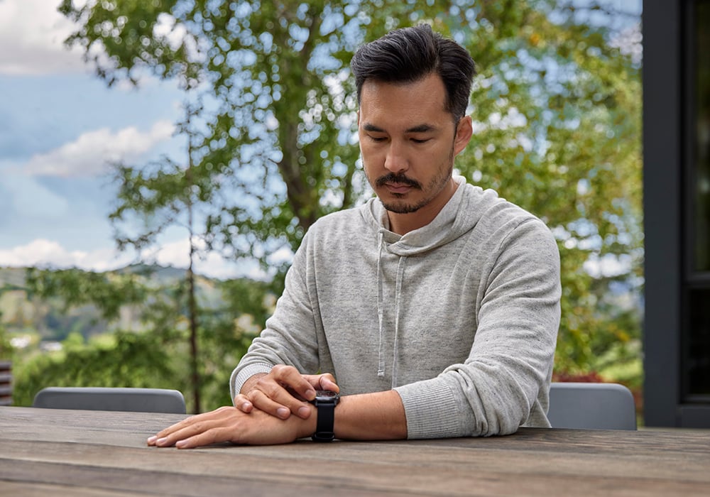 A man sitting at a table outside while looking down at his wearable. A landscape of trees is behind him.